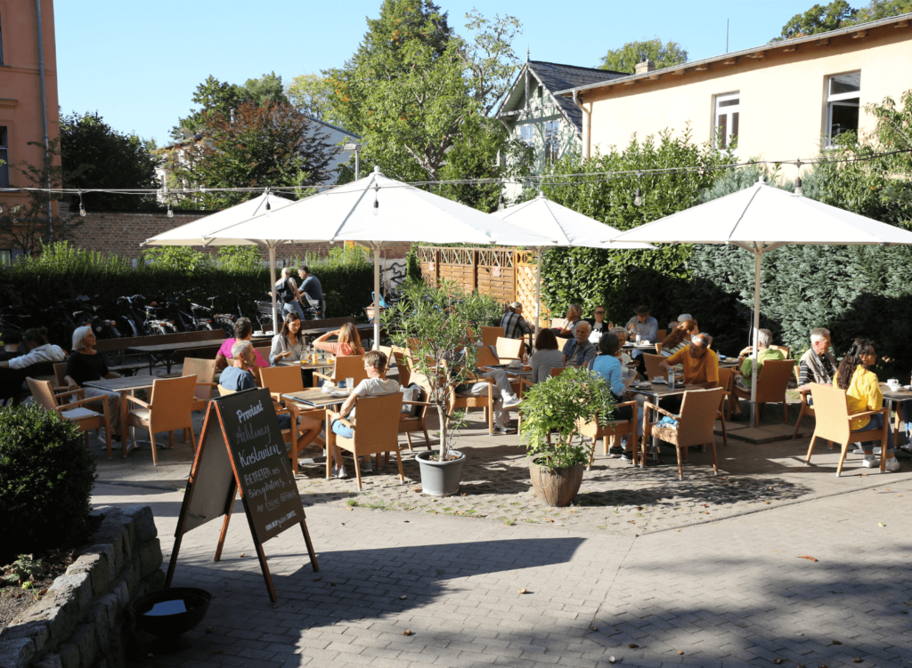 Blick auf die gefühlte Terrasse. Es sitzen Menschen an Tischen und essen Eis und Kuchen. Es ist Sommer